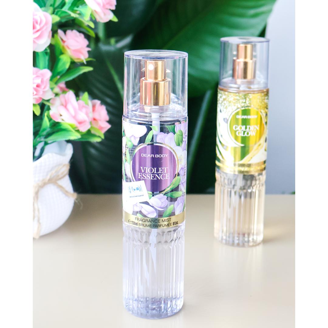 Two clear glass perfume spray bottles with gold atomizers on a light beige surface. The front bottle is labeled 'Ding Dong Body Violet Essence' with purple and white floral graphics, and the rear bottle is labeled 'Green Tea Blossom' with yellow and green graphics.