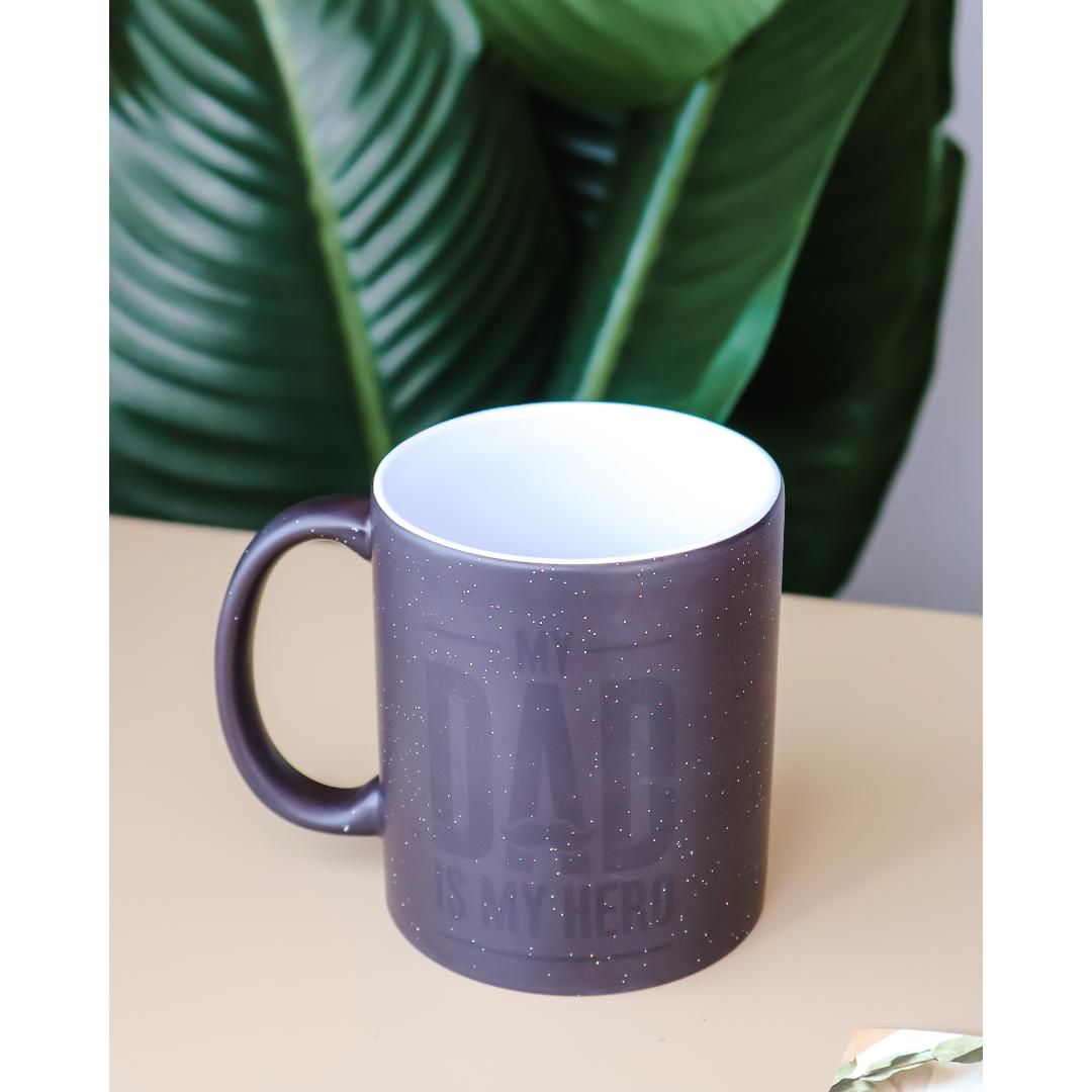 A dark gray speckled ceramic mug with a white interior and handle, featuring the embossed text 'THE DAD IS MY HERO' on the front.