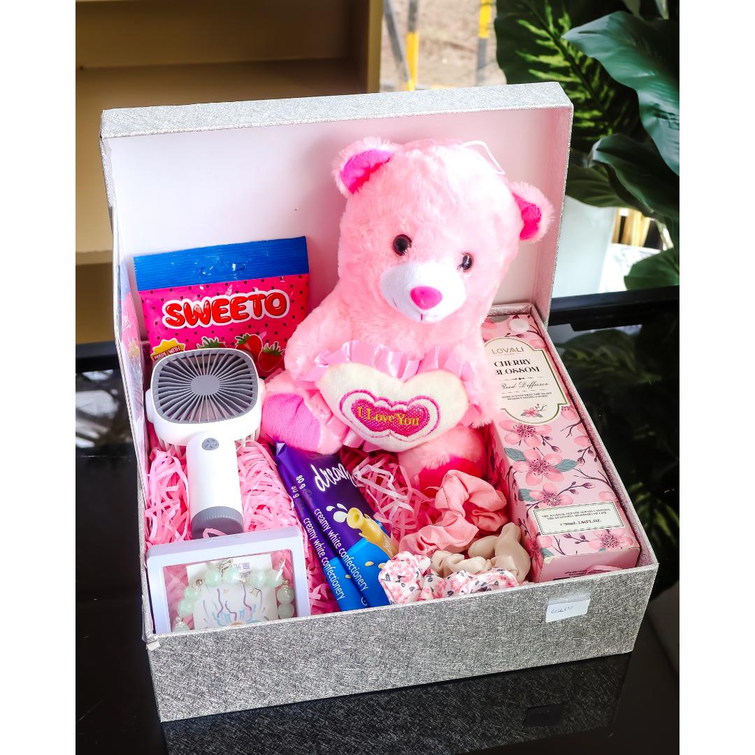 A birthday gift box filled with pink-themed items, including a pink teddy bear holding a heart, a small pink handheld fan, a Sweetto candy bag, a chocolate bar, and pink accessories, set on a dark reflective surface.