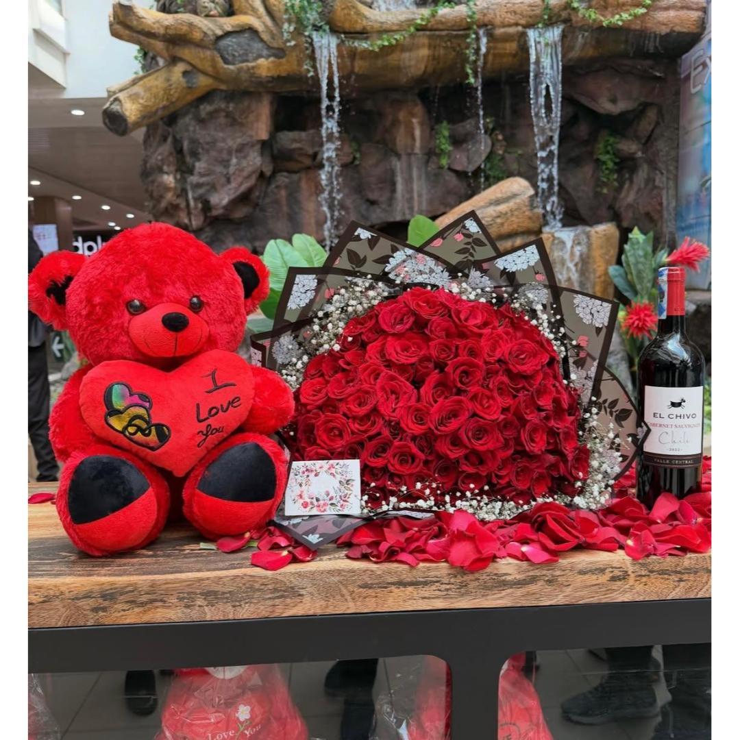 A red plush teddy bear with 'I Love you' embroidered on its chest sits beside a large bouquet of red roses, and a bottle of red wine