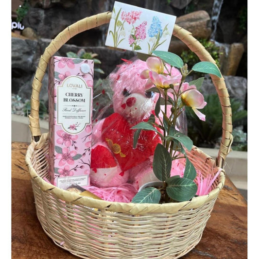 A wicker gift basket arranged on a wooden surface with a pink plush teddy bear in a red outfit, a pink floral home reed diffuser, and a small card.