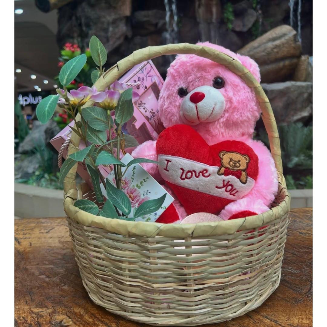 A pink plush teddy bear gift set in a woven wicker basket, featuring a red heart-shaped pillow that says 'I love you' and pink flower stems.