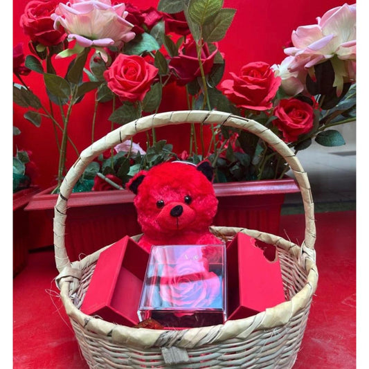 A wicker gift basket filled with a small red plush teddy bear, red gift boxes, and a clear box containing a red rose.