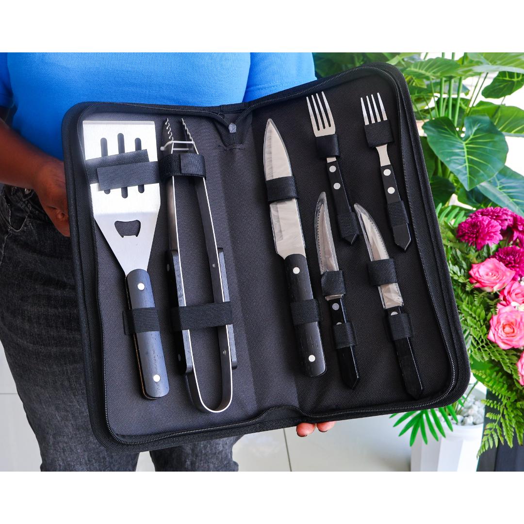 A 7-piece barbecue tool set with stainless steel blades and black handles, displayed inside an open black zippered carrying case. The set includes a spatula, tongs, three knives, and two forks.