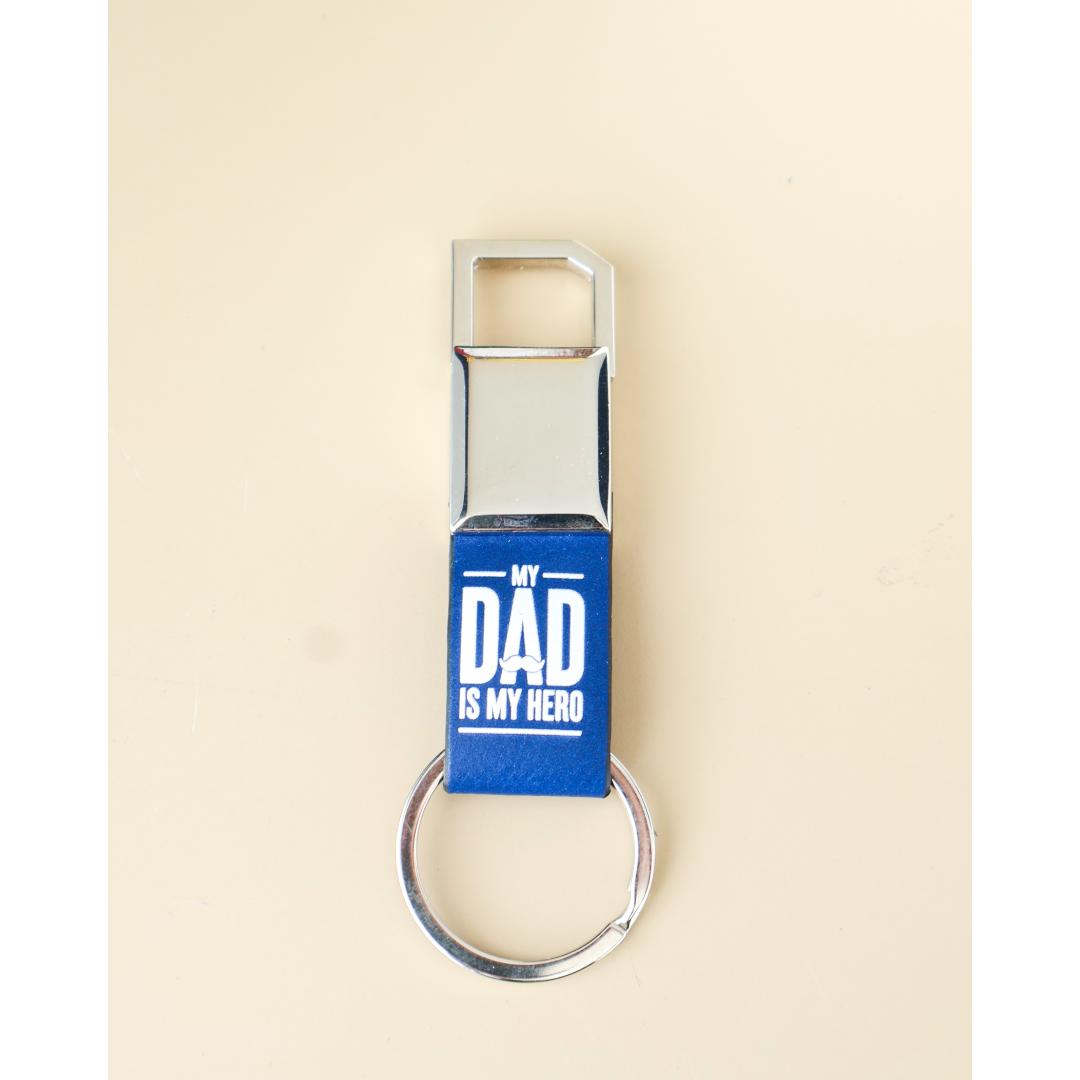 A bottle opener keychain with a polished silver metal body and a blue leather wrap featuring the white text 'MY DAD IS MY HERO', set against a solid cream background.