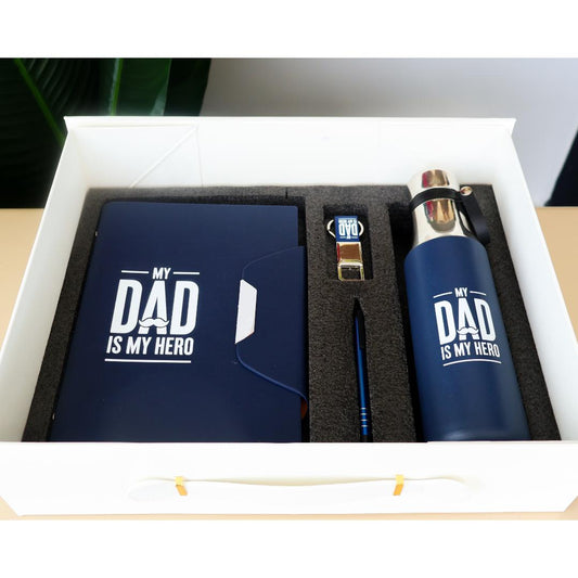 A boxed gift set for Dad featuring a navy blue notebook, a matching insulated bottle, a small keychain, and a pen, all printed with the white text 'MY DAD IS MY HERO'. 