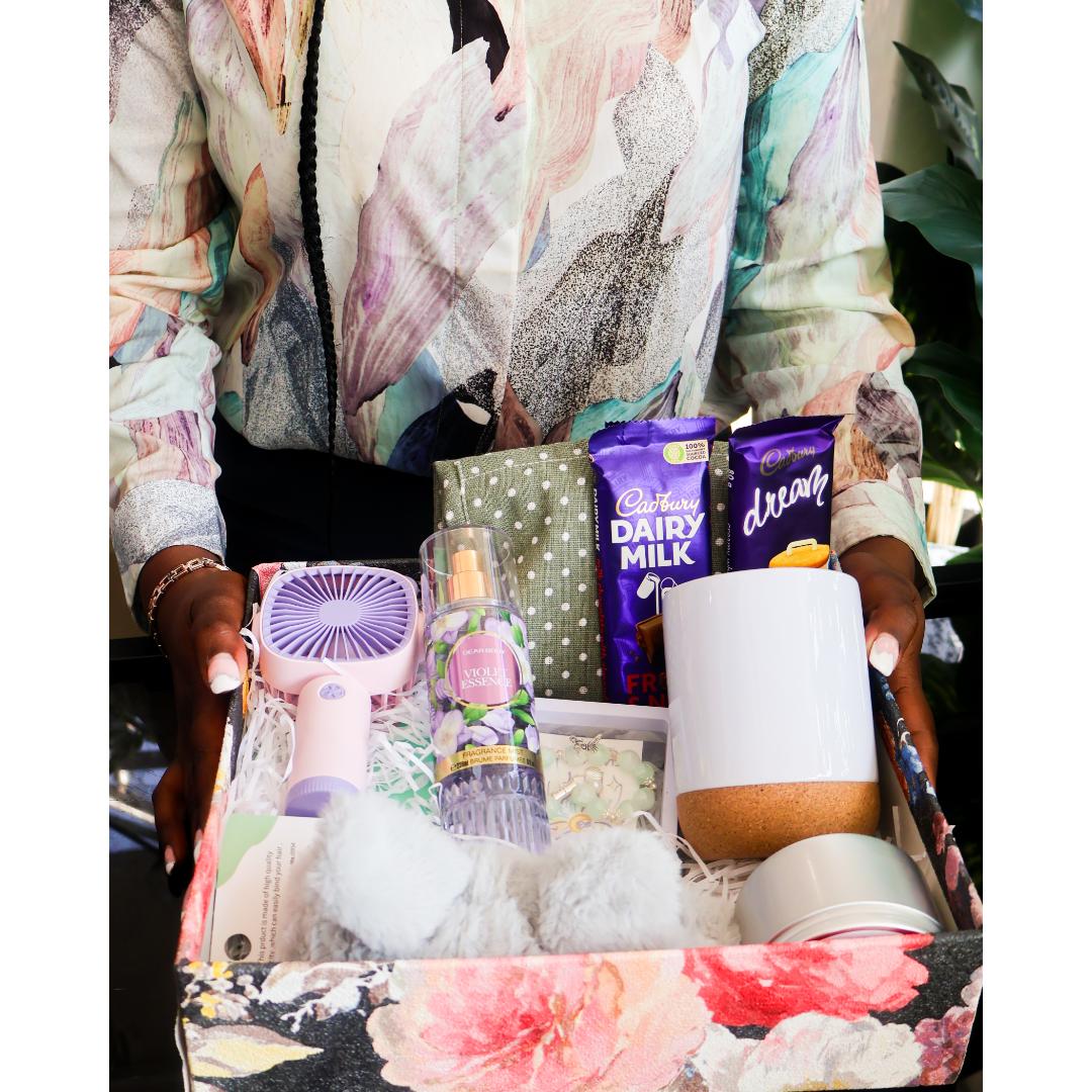 A gift box filled with assorted items including a small lavender fan, a bottle of body spray, a white mug with a cork base, plush toys, a scented candle, and chocolate bars.