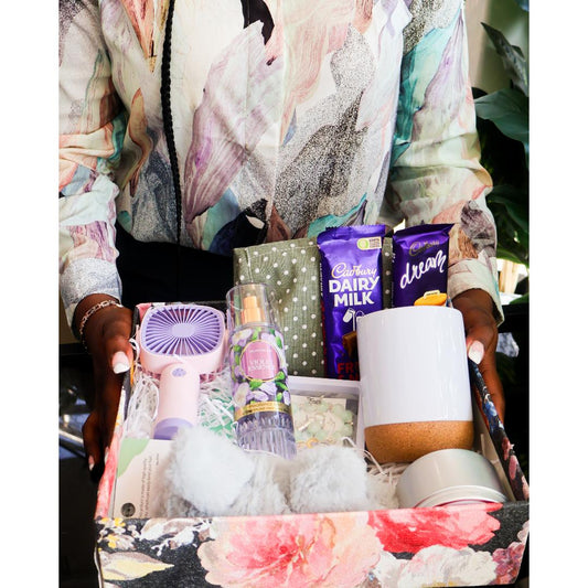 A gift box filled with assorted items including a small lavender fan, a bottle of body spray, a white mug with a cork base, plush toys, a scented candle, and chocolate bars.