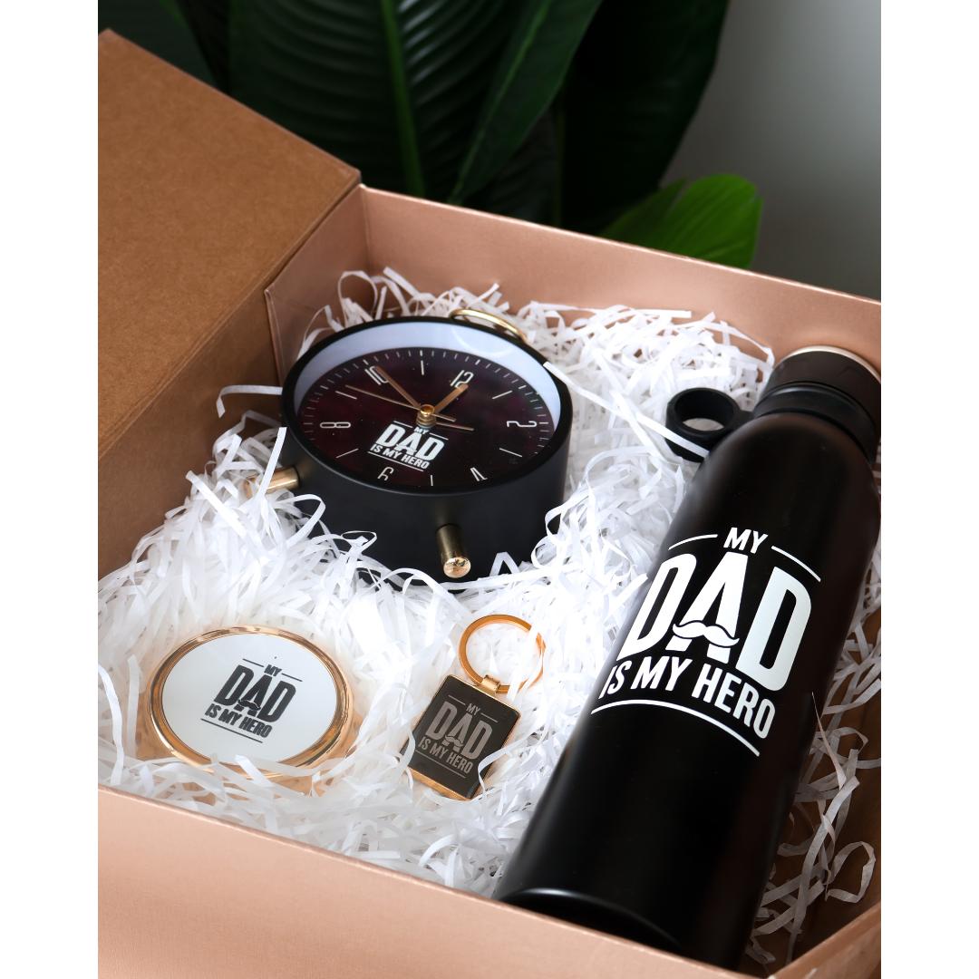 A Dad-themed gift set, featuring a black analog clock with gold details, a black insulated bottle with white text reading 'MY DAD IS MY HERO', a round tin with a lid that says 'DAD DAY 1', and a small keychain.