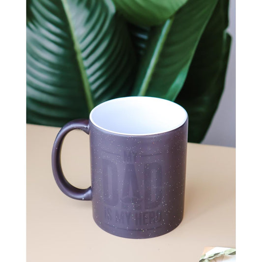 A dark gray speckled ceramic mug with a white interior and handle, featuring the embossed text 'THE DAD IS MY HERO' on the front.