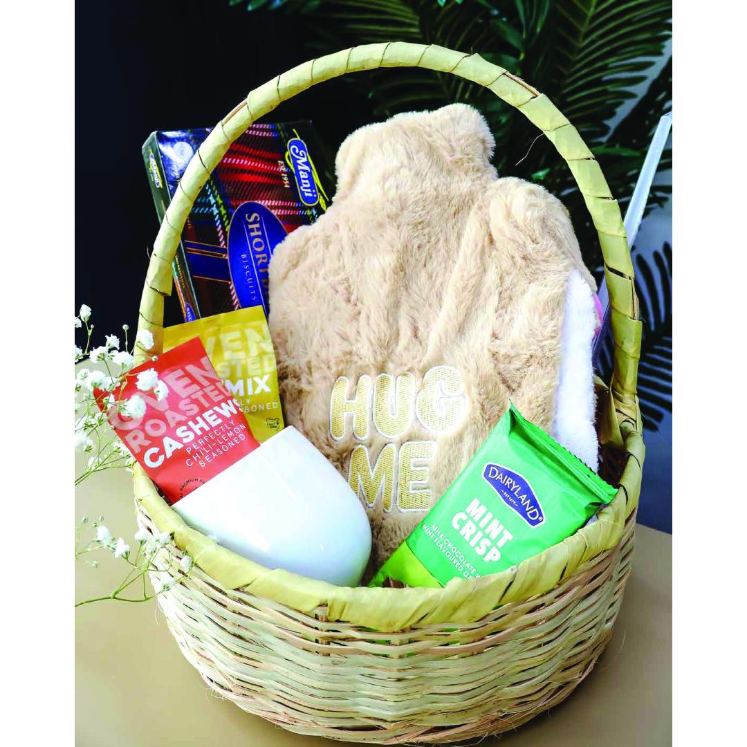A woven wicker gift basket filled with assorted treats and comfort items, including a beige plush hot water bottle, a white ceramic mug, a green packet of Dairymaid Mint Crisp, a red packet of Rosenzweig Cashew Mix, and a blue box of biscuits.