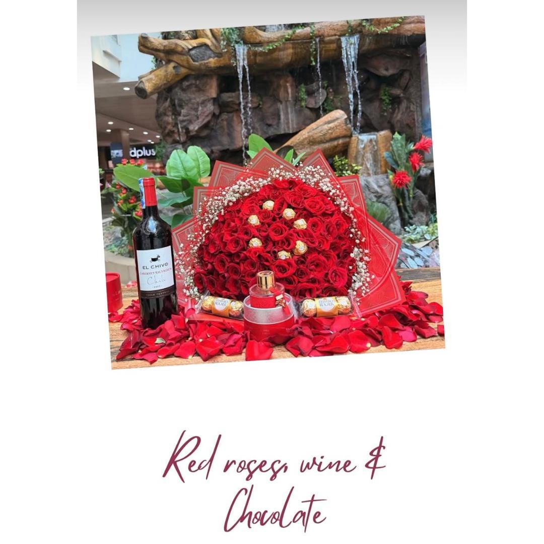 A red rose bouquet arrangement with a bottle of red wine and several chocolate-covered treats.