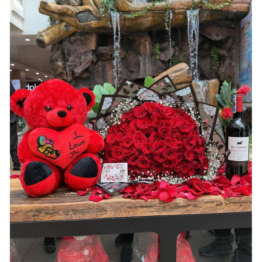 A red plush teddy bear with 'I Love you' embroidered on its chest sits beside a large bouquet of red roses, and a bottle of red wine