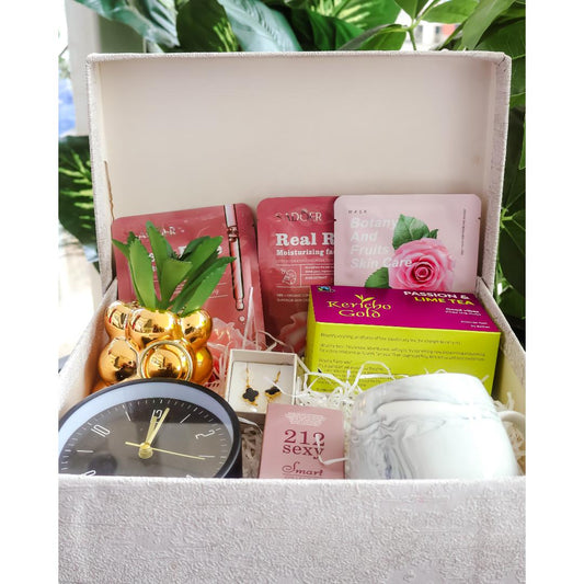 A curated gift box filled with beauty and lifestyle items, including pink rose face masks, passion and lime tea box, a small black clock, gold decorative ornaments, and a white marble mug.