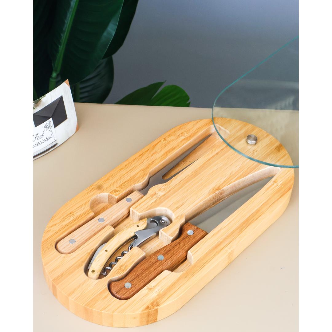 A bamboo wine and cheese spreader set on an oval wooden tray, shown with a glass lid partially lifted on a tabletop. The set includes a corkscrew, cheese knife, and spreader fork, with visible wood and metal details.