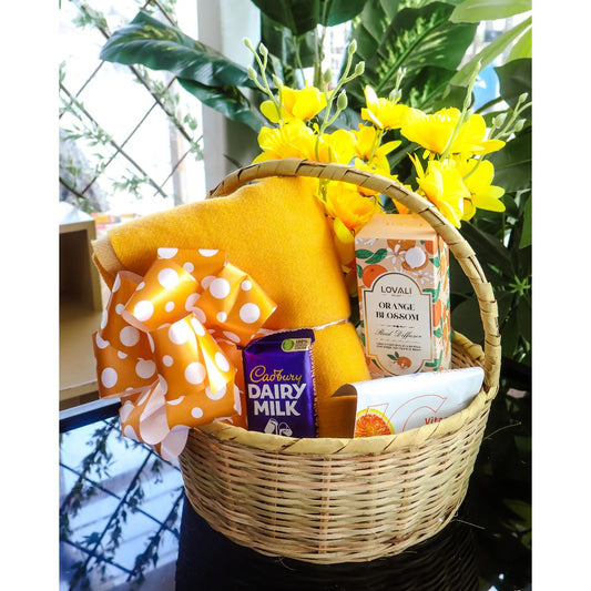 A wicker gift basket filled with a yellow scarf, an orange blossom home reed diffuser, a Cadbury Dairy Milk chocolate bar, and a face mask.